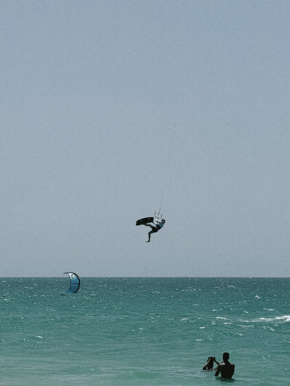 Mid-jump over the kiters' break, a swimmer's head bobbing in the foreground
