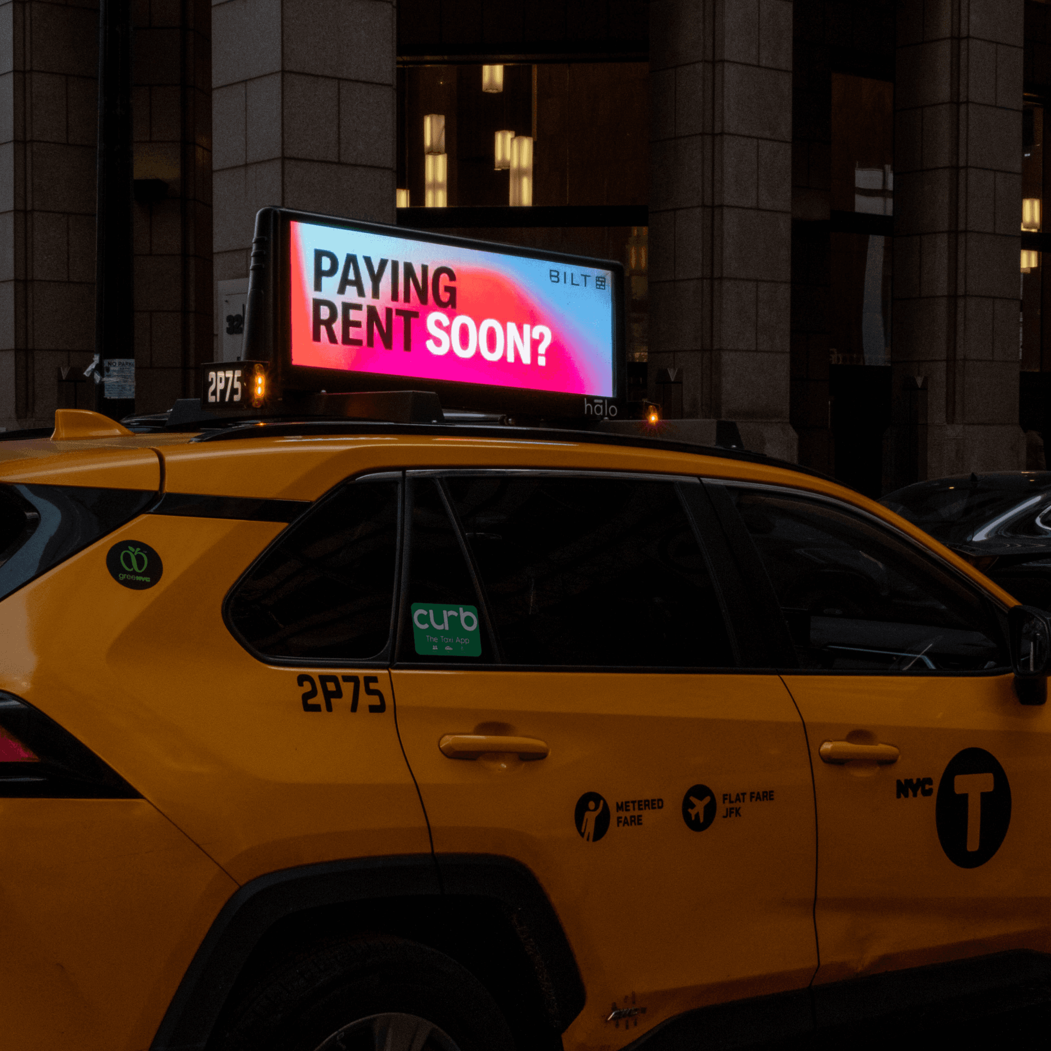 NYC yellow taxi at night with a Bilt roof-top display reading 'Paying Rent Soon?' in pink and white type